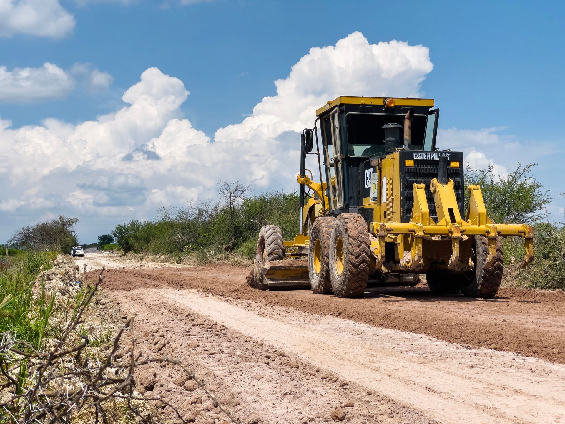 Yellow road grader leveling a dirt road in rural area on a sunny day.